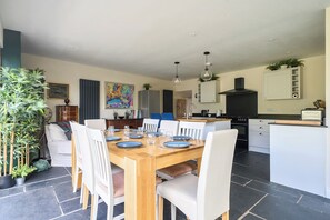 A modern kitchen with a central island, white cabinetry, and a dining area with a wooden table and chairs. - 4-bed Cottage - Pets With Garden & Wood Burner (Helston)