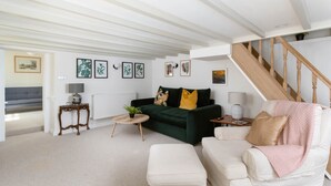 A living room with a green sofa, a white armchair, a small round table, and a staircase. - 4-bed Cottage - Pets With Garden & Wood Burner (Helston)