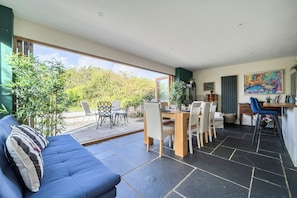 A modern living room with a blue sofa, dining area with chairs and a table, and a view of the outdoors through large windows.