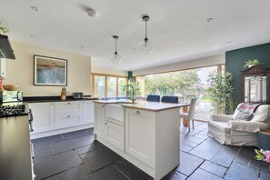 A modern kitchen with white cabinets, a central island, and a dining area with a view of the garden. - 4-bed Cottage - Pets With Garden & Wood Burner (Helston)