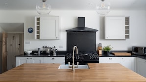 A modern kitchen with a central island, black stove, white cabinets, and pendant lighting. - 4-bed Cottage - Pets With Garden & Wood Burner (Helston)