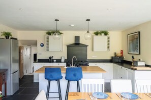 A modern kitchen with a central island, bar stools, a dining table, and various appliances. - 4-bed Cottage - Pets With Garden & Wood Burner (Helston)