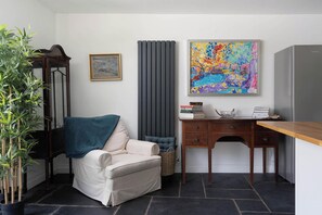 Ein Zimmer mit einem weißen Sessel, einem Schreibtisch mit Büchern und einer Schale, einem Heizkörper, einem farbenfrohen abstrakten Gemälde und einer Topfpflanze.