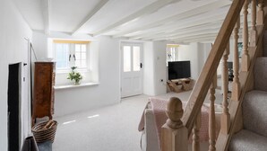 A spacious living room with a high ceiling, a wooden staircase, a television, and a window with a view. - 4-bed Cottage - Pets With Garden & Wood Burner (Helston)