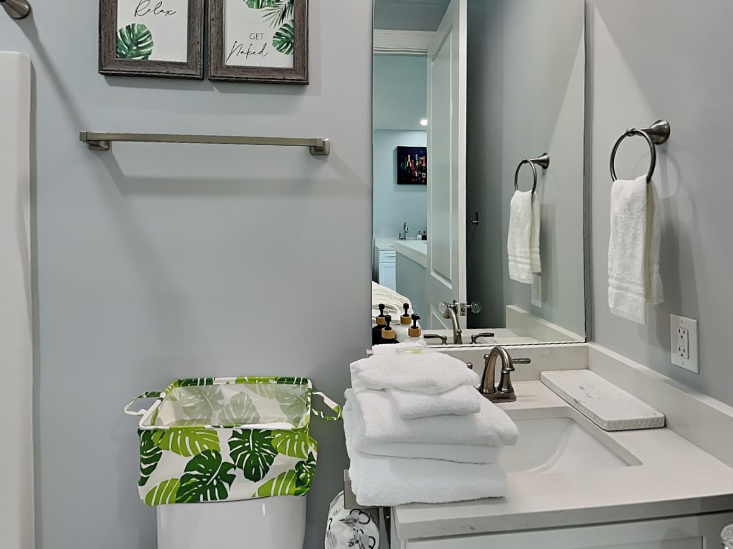 A bathroom with a white sink, a mirror, and a towel rack. There are two framed pictures on the wall and a green plant-patterned basket on the floor.