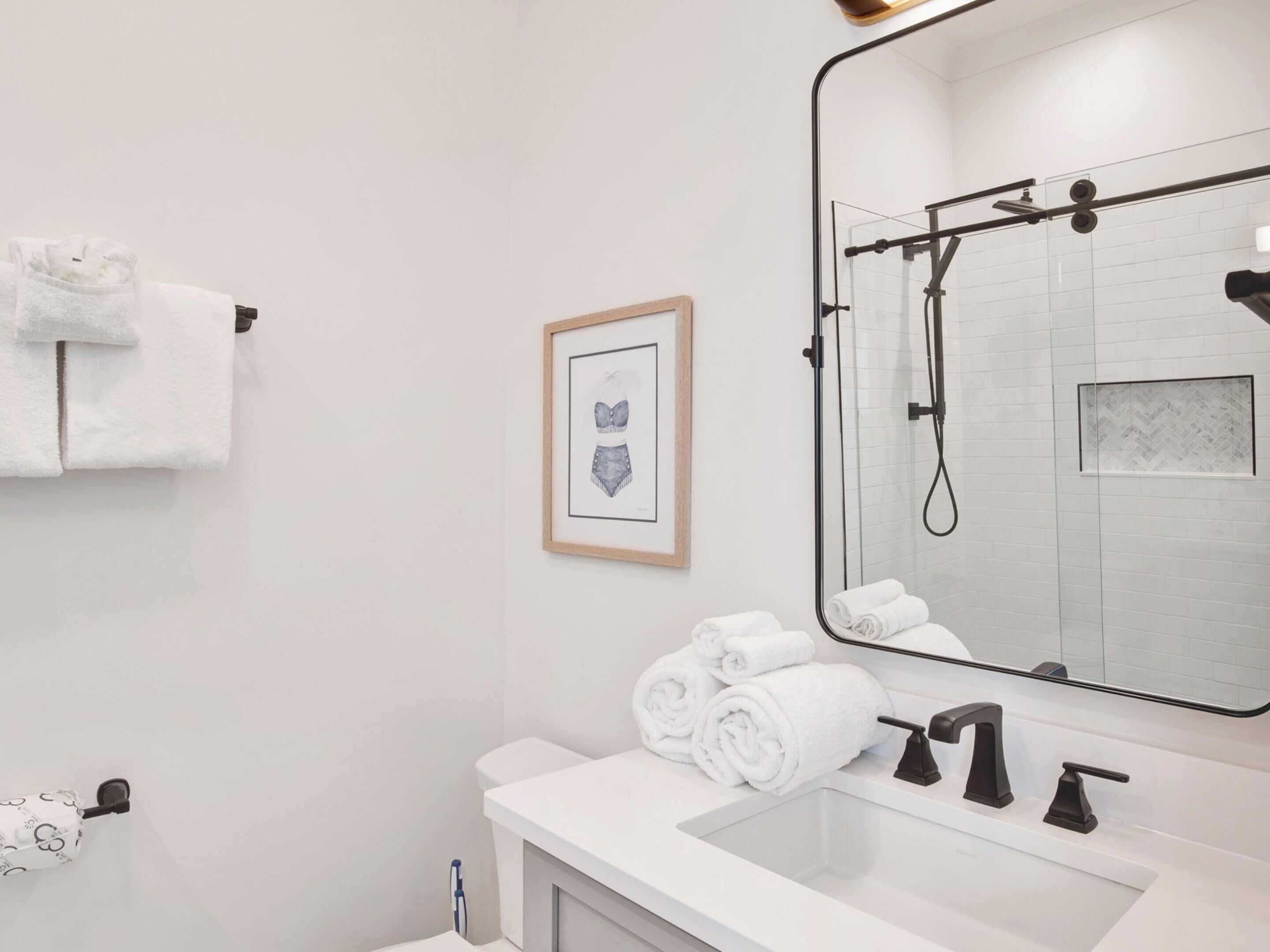 A modern bathroom with a white sink, black faucets, and a large mirror. There are rolled towels on the sink and a framed picture on the wall.