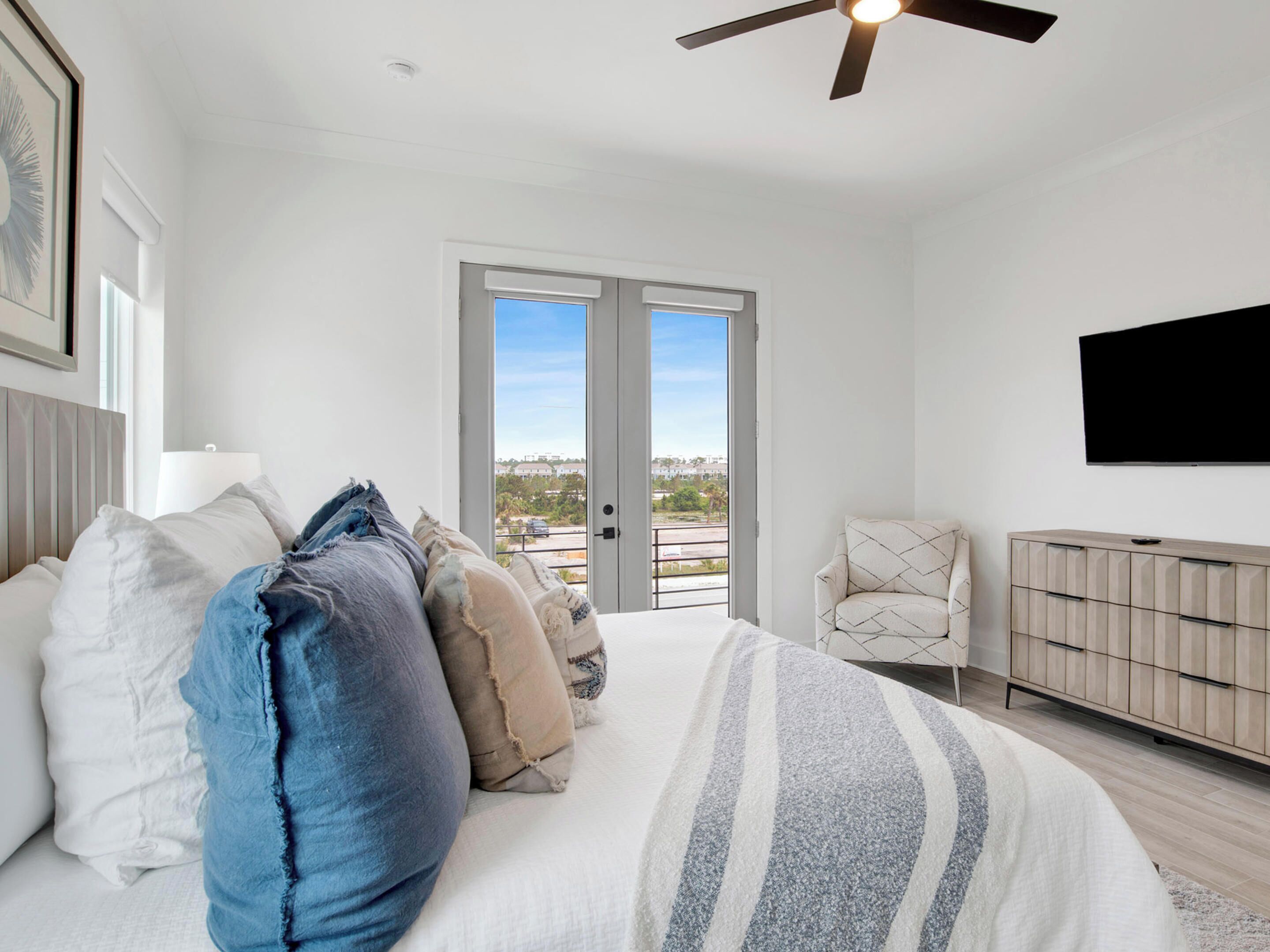 A bedroom with a bed, pillows, a chair, a TV, and a door leading to a balcony with a city view.