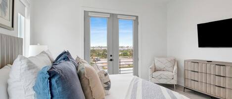 A bedroom with a bed, pillows, a chair, a TV, and a door leading to a balcony with a city view.