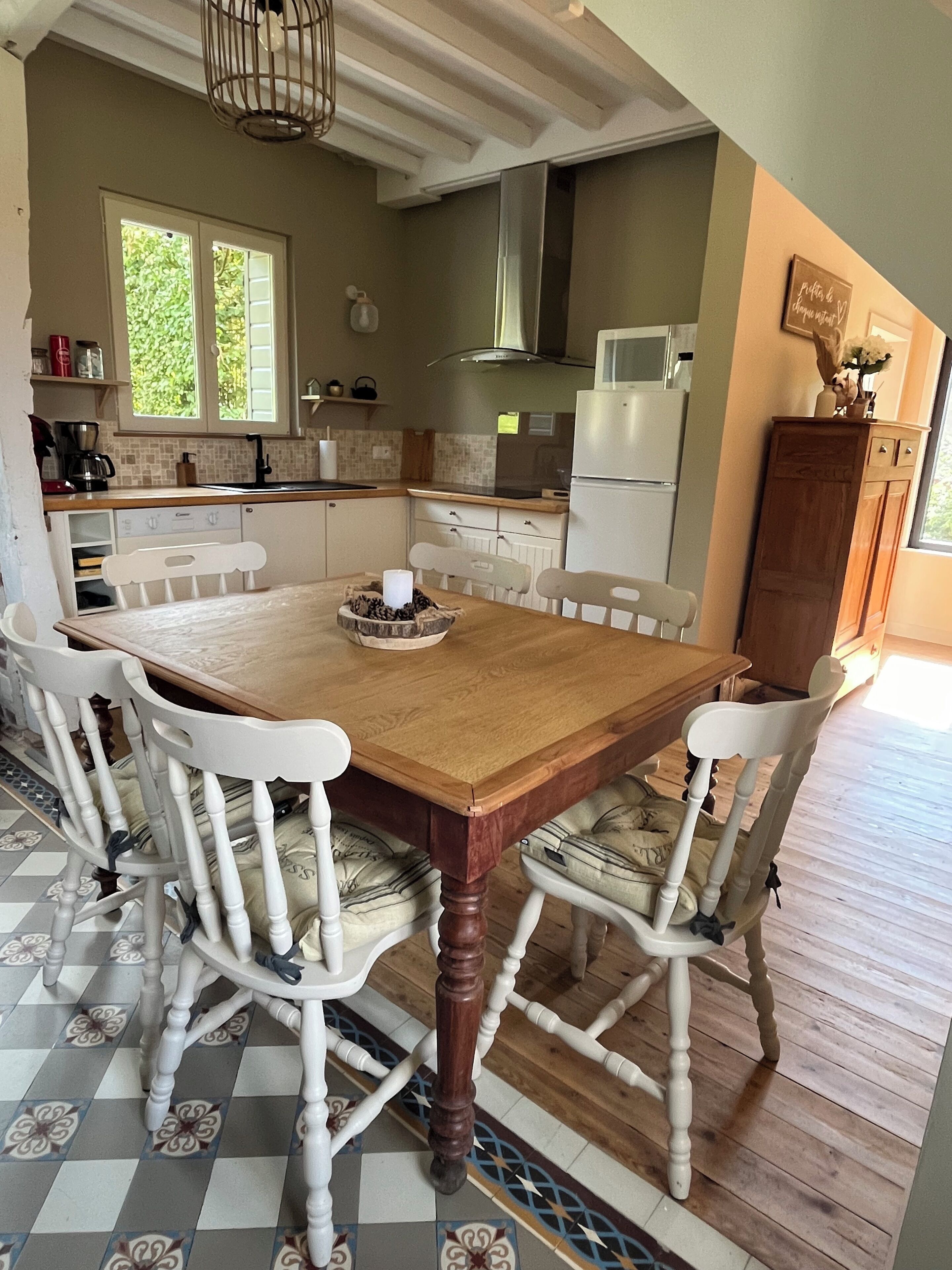 Un coin repas avec une table en bois et des chaises blanches, une cuisine en arrière-plan et un sol à motifs.