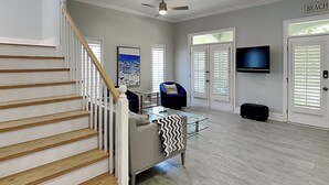 Un vaste espace de vie comprenant un escalier, un ventilateur de plafond, un téléviseur et une porte portant l’inscription « PLAGE ».