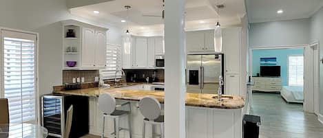 Una cocina moderna con armarios blancos, encimera de granito y electrodomésticos de acero inoxidable. Hay tres taburetes de bar y una mesa de comedor de vidrio.