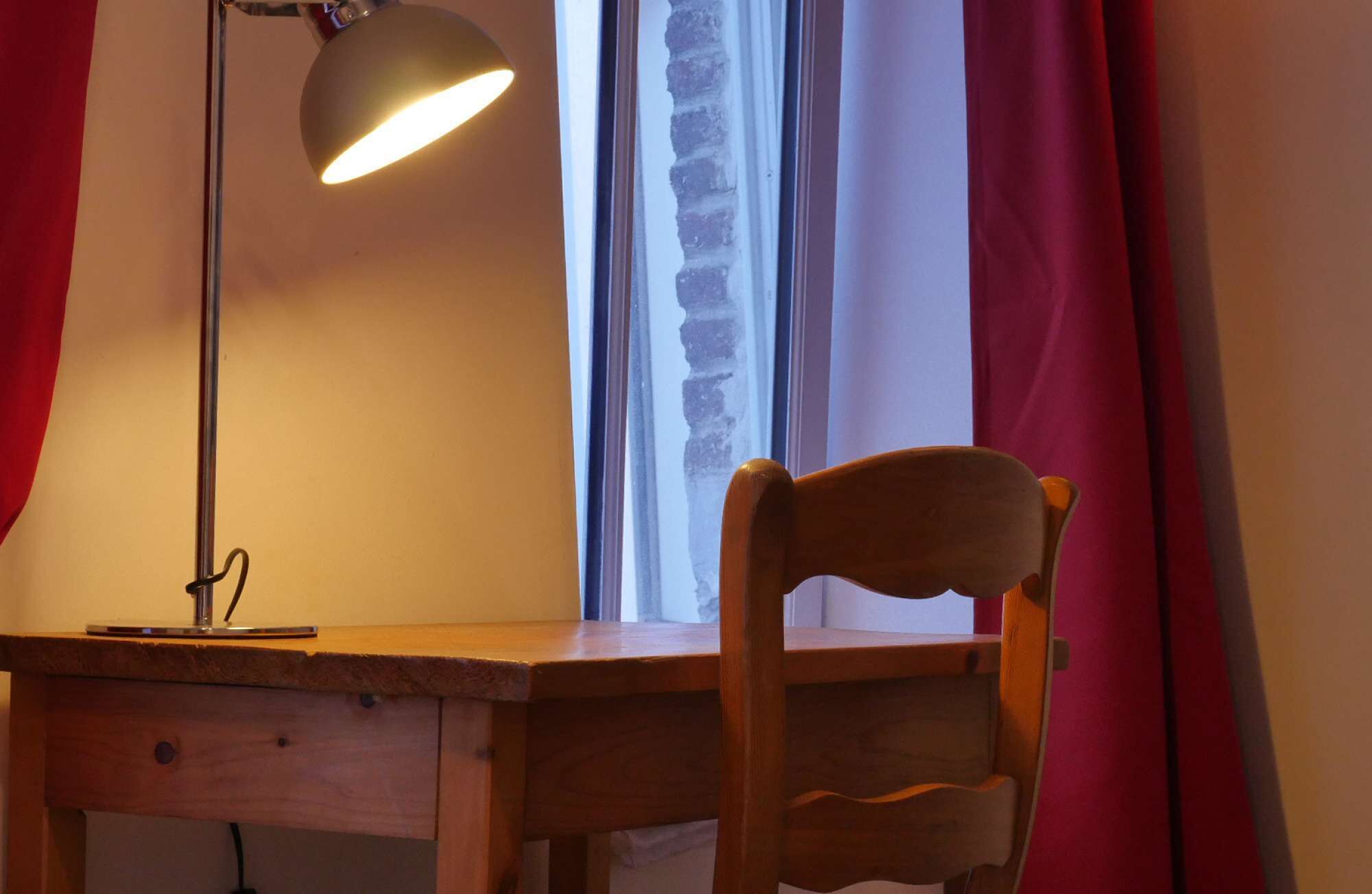Ett skrivbord i trä med en lampa, en stol och ett fönster med gardiner.