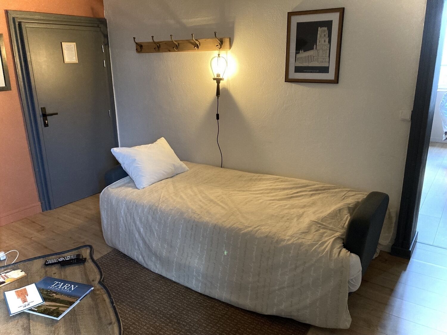 A single bed with a white pillow and a light-colored blanket in a room with a wooden headboard, a wall-mounted light, and a framed picture.