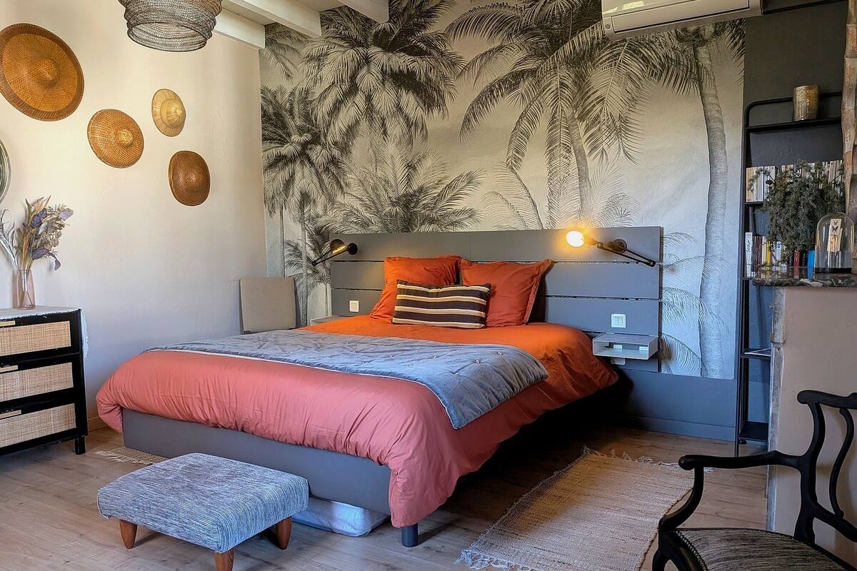 A bedroom with a bed, bedside tables, a nightstand, a chair, and a wall with a tropical mural.