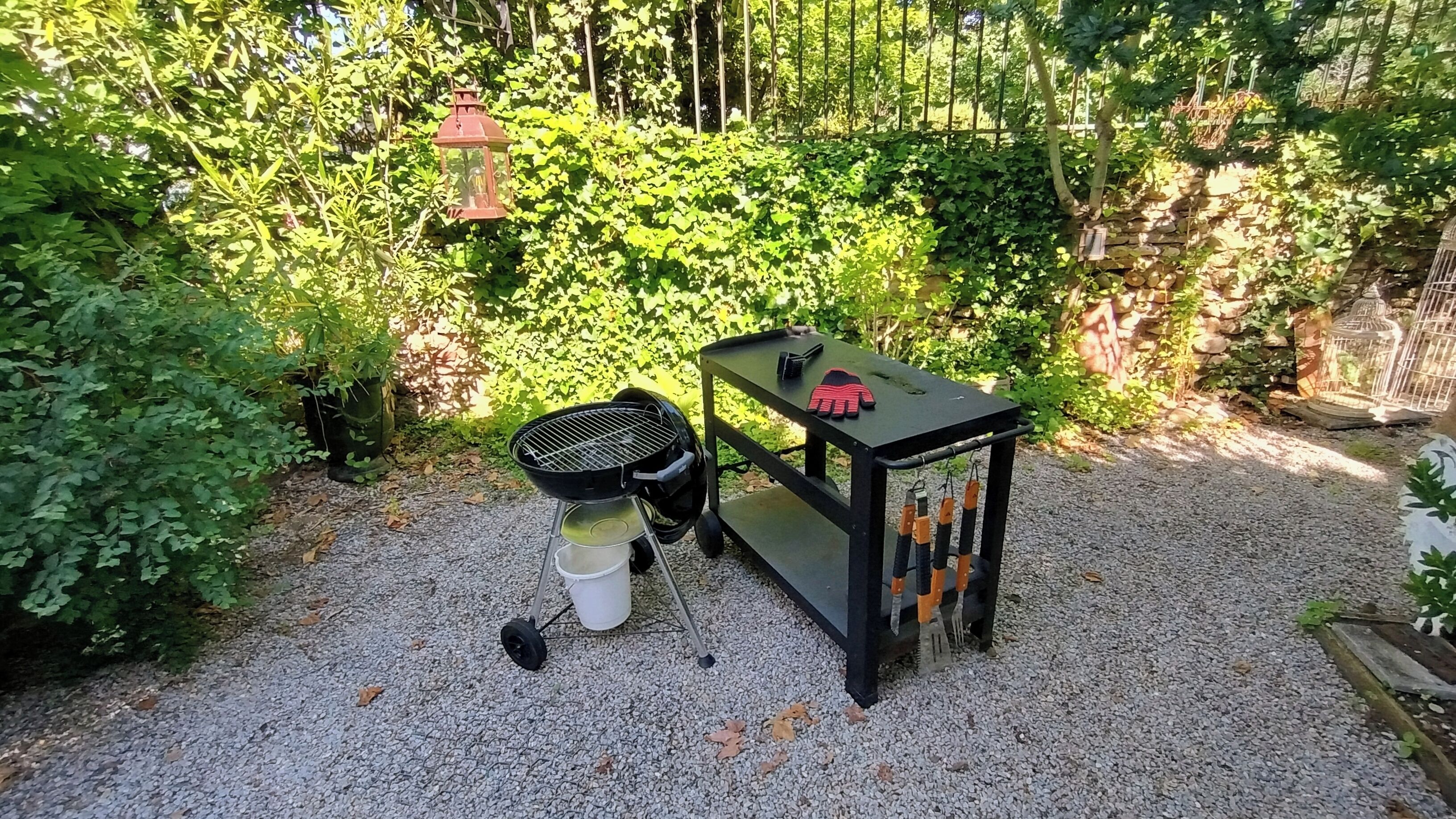 Een terras met grind, een barbecue, een witte emmer en een zwarte kar met tuingereedschap.