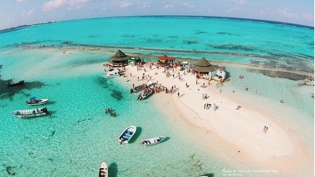 An aerial view of a beach with clear turquoise waters, several boats, and a gathering of people near structures with thatched roofs.