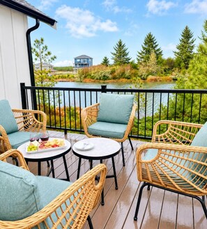 Un balcon agrémenté de meubles en osier, une petite table sur laquelle est disposée une assiette de fruits, et une vue sur un lac et des arbres.