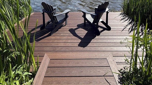 Een houten terras met twee Adirondack stoelen gericht op een kalm meer, omringd door groen en bomen.