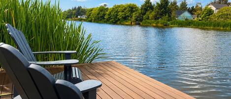 A wooden deck with Adirondack chairs overlooking a calm lake, surrounded by lush greenery and houses.