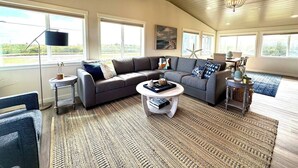 A spacious living room with a large sectional sofa, a round coffee table, and a chandelier. There are multiple windows providing natural light and a view of the outdoors. - Waterfront Duplex w/Dock | Near Damon Point Beach (Ocean Shores)