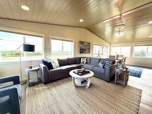 A spacious living room with a large sectional sofa, a round coffee table, and a chandelier. There are multiple windows providing natural light and a view of the outdoors. - Waterfront Duplex w/Dock | Near Damon Point Beach (Ocean Shores)