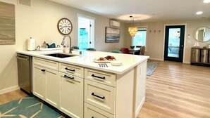 A modern kitchen with a central island, a dining area with a chandelier, and a view of the outdoors through a door.
