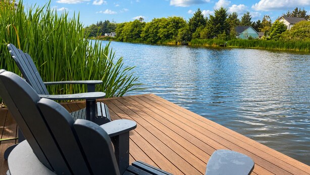 A wooden deck with Adirondack chairs overlooking a calm lake, surrounded by lush greenery and houses.