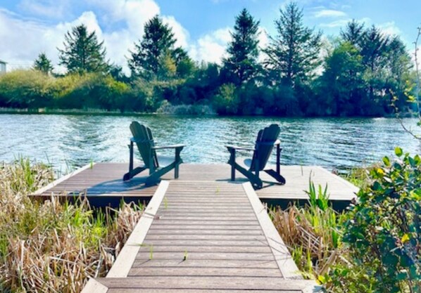 A wooden dock with two Adirondack chairs facing a calm lake, surrounded by dense trees and greenery.