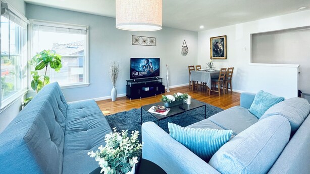 Ein modernes Wohnzimmer mit einer grauen Sofagarnitur, einem Couchtisch, einem Fernseher und einem Essbereich mit Tisch und Stühlen.