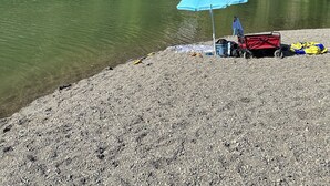 Un cadre idyllique au bord d’un lac, avec une plage de galets, un parasol bleu et une petite charrette rouge.