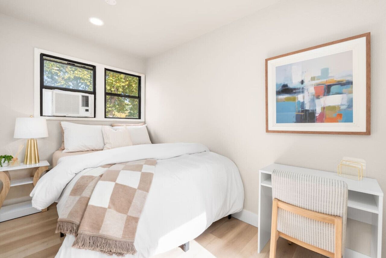 A modern bedroom with a large bed, two windows with air conditioning, a bedside table with a lamp, and a framed abstract painting on the wall.