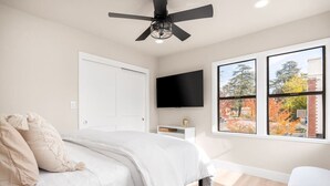 Ein Schlafzimmer mit einem Bett, einem Deckenventilator, einem an der Wand befestigten Fernseher und einem Fenster mit Blick auf Bäume.