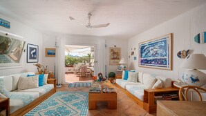 Ein gut beleuchtetes Wohnzimmer mit zwei weißen Sofas, einem Couchtisch und einem Deckenventilator. An den Wänden befinden sich gerahmte Bilder und es gibt einen Blick auf die Außenpatiofläche.