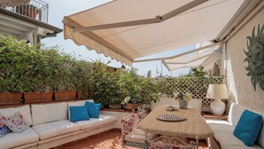 Eine Terrasse im Freien mit einem weißen Sofa, einem Tisch und Stühlen. Übertöpfe mit Pflanzen und einem dekorativen Wandelement.