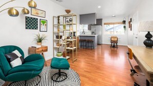 Un salon moderne avec un fauteuil turquoise, une table basse en bois et un tapis noir et blanc. On aperçoit, au fond, une cuisine avec un plan de travail gris et une fenêtre.