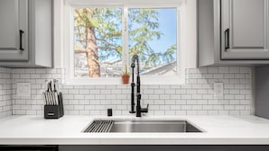Une cuisine avec un mur recouvert de carreaux blancs, une fenêtre donnant sur des arbres et un robinet noir.