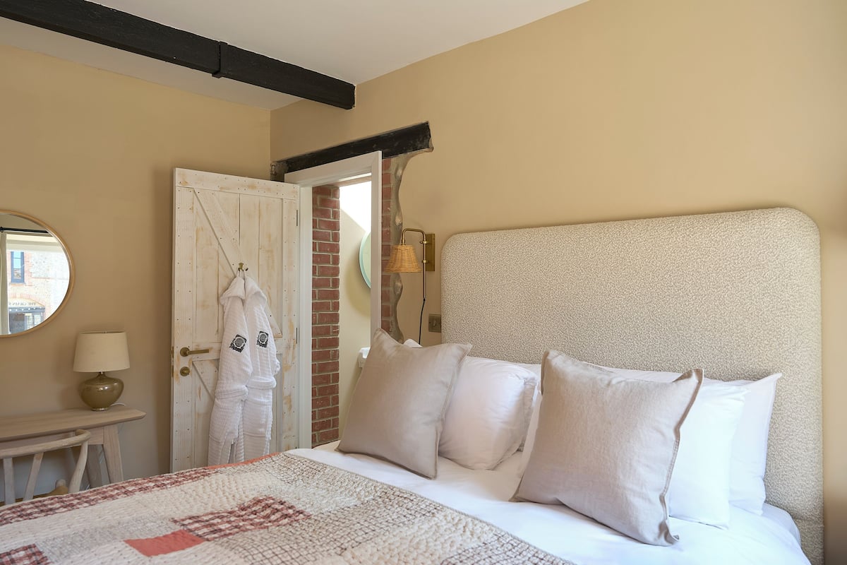 Un dormitorio con una cama grande, ropa de cama blanca, un espejo redondo, una mesita de noche con una lámpara y una puerta que da a una pared de ladrillo.