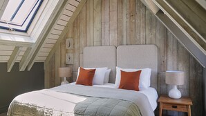 A bedroom with a sloped ceiling, wooden walls, a bed with white and brown bedding, two bedside tables with lamps, and a skylight. - The Maltings (Holt)