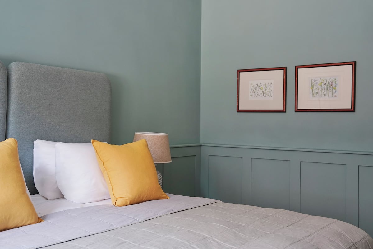 Una cama bien hecha con cabecera gris, almohadas blancas y amarillas, y colcha gris. Hay dos láminas botánicas enmarcadas en la pared y una lámpara de noche.