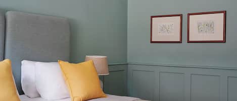 Una cama bien hecha con cabecera gris, almohadas blancas y amarillas, y colcha gris. Hay dos láminas botánicas enmarcadas en la pared y una lámpara de noche.