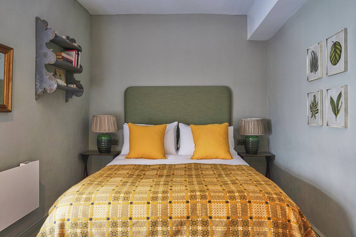 Un dormitorio con cama, dos lámparas de noche, cabecero, estante con libros e impresiones botánicas enmarcadas en la pared.