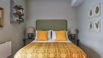 A bedroom with a bed, two bedside lamps, a headboard, a shelf with books, and framed botanical prints on the wall.
