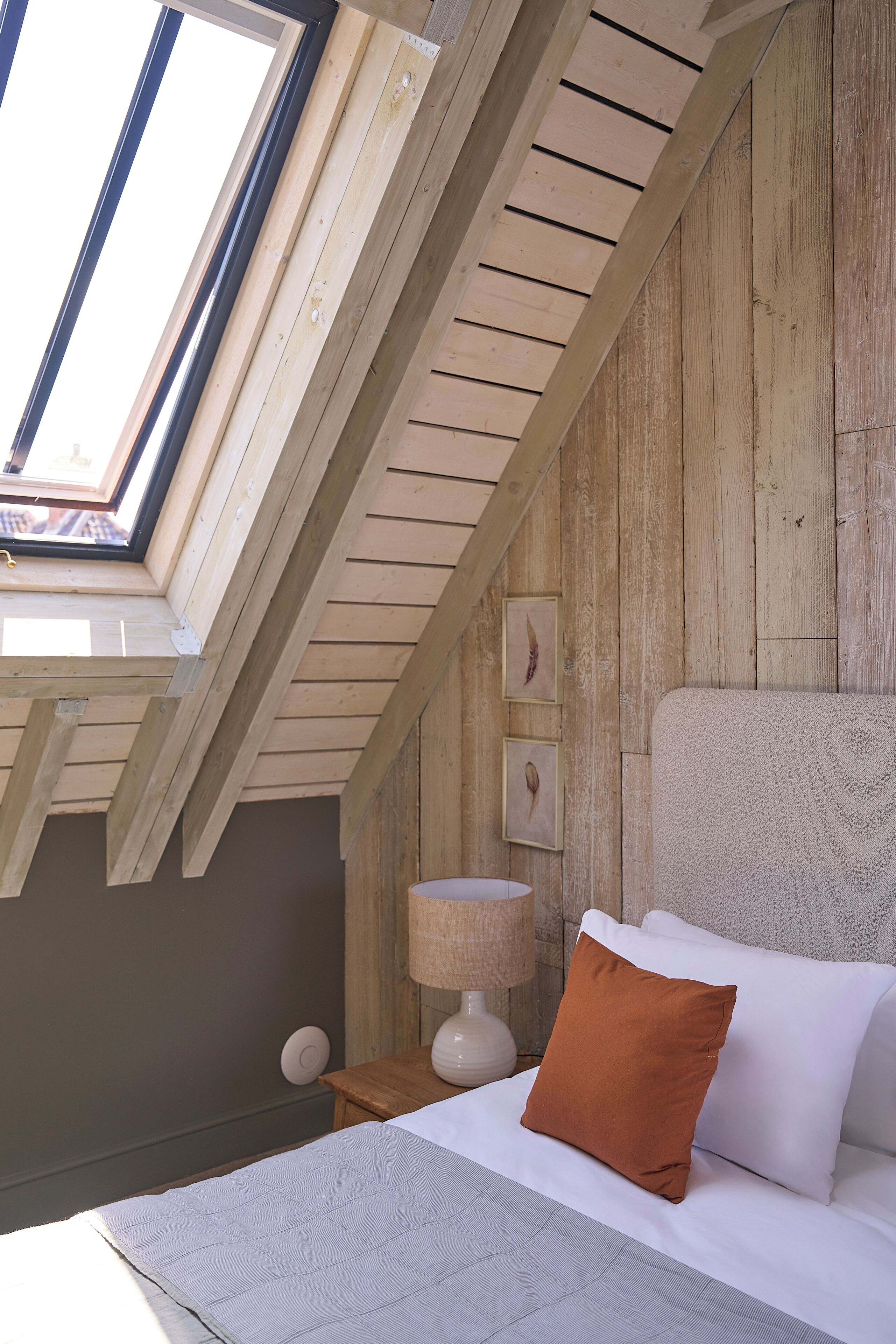 Ein Schlafzimmer mit schrägen Wänden, einem Oberlicht, einem Bett mit weißen und braunen Kissen, einer Nachttischlampe und Holzwänden.