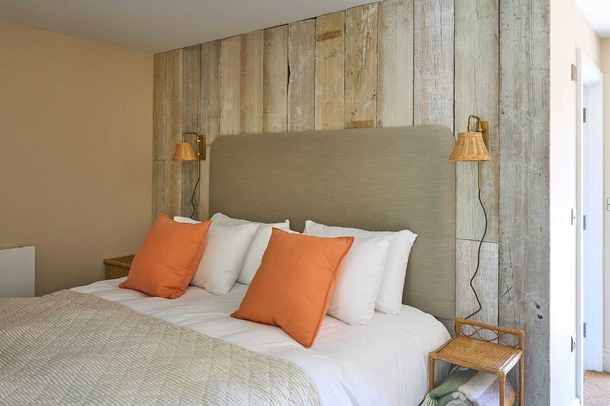 Un dormitorio con cabecera de madera, cama con ropa de cama blanca y naranja, dos lámparas de noche y una silla de mimbre.