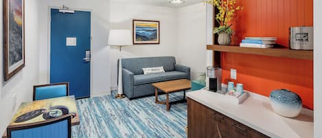 A room with a blue door, a sofa, a small table, a coffee station, and a red wall with a shelf.