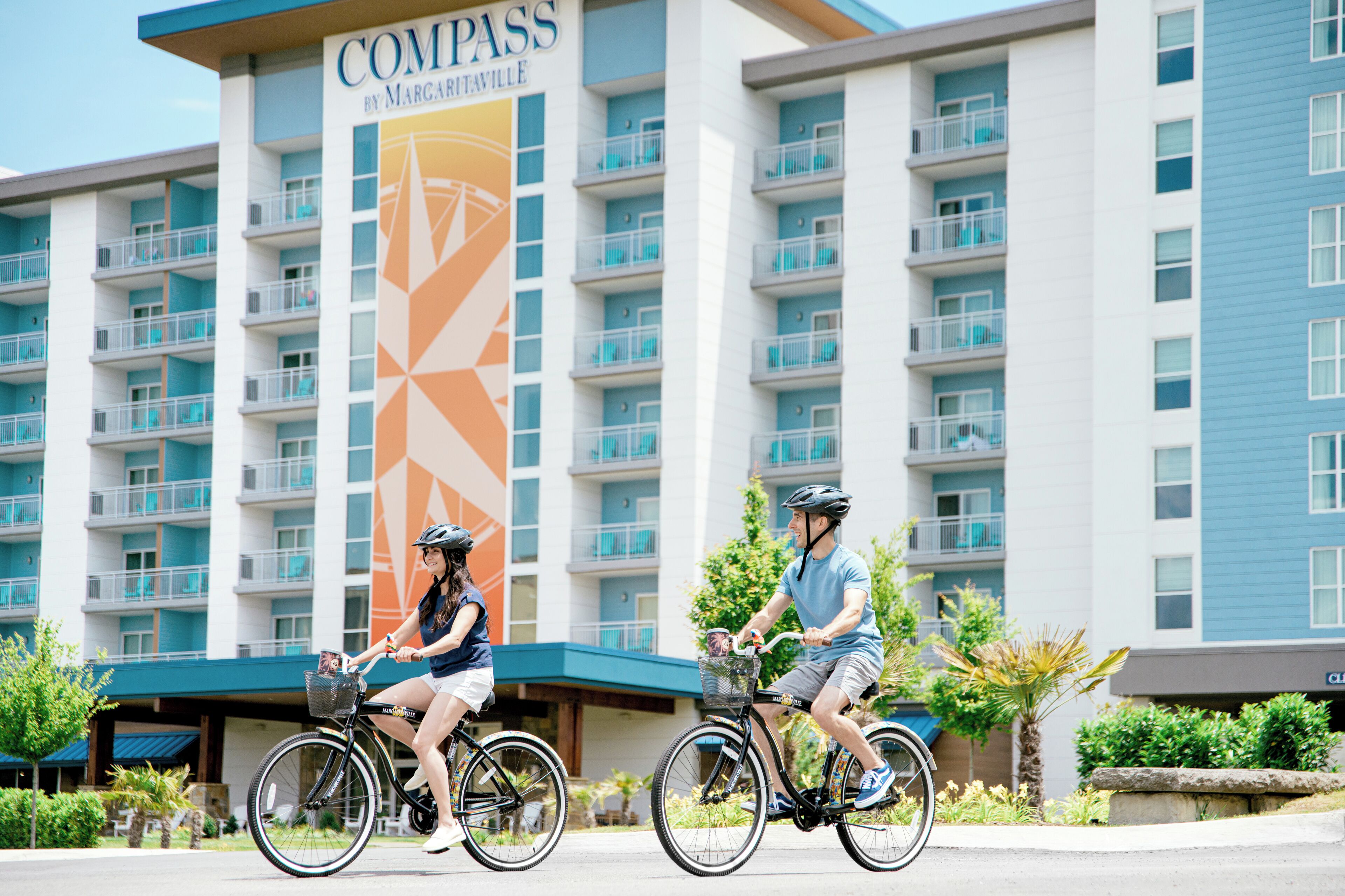 Two people riding bicycles in front of a hotel with a large sign reading "Compass by Margareville."