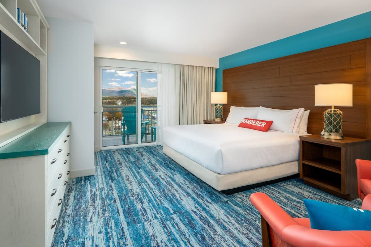 A hotel room with a large bed, a desk, a chair, and a view of mountains through the balcony doors.