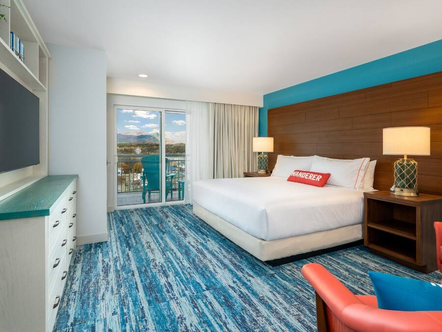 A hotel room with a large bed, a desk, a chair, and a view of mountains through the balcony doors.