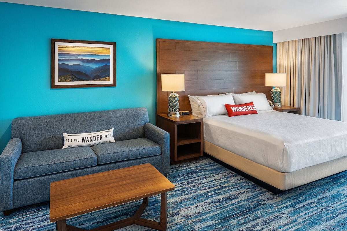 A hotel room with a blue wall, a bed with white bedding, a grey sofa, a wooden coffee table, and a painting of a mountain landscape.