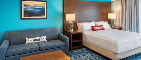 A hotel room with a blue wall, a bed with white bedding, a grey sofa, a wooden coffee table, and a painting of a mountain landscape.